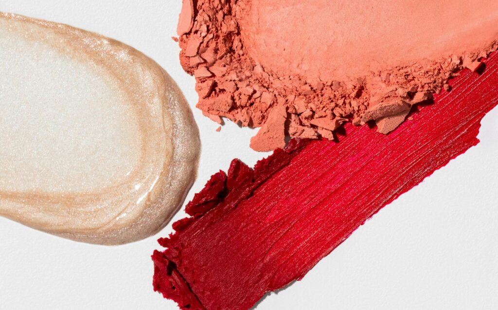 Red lipstick, peach blush powder and pearlescent texture representing lanolin in cosmetic formulations.