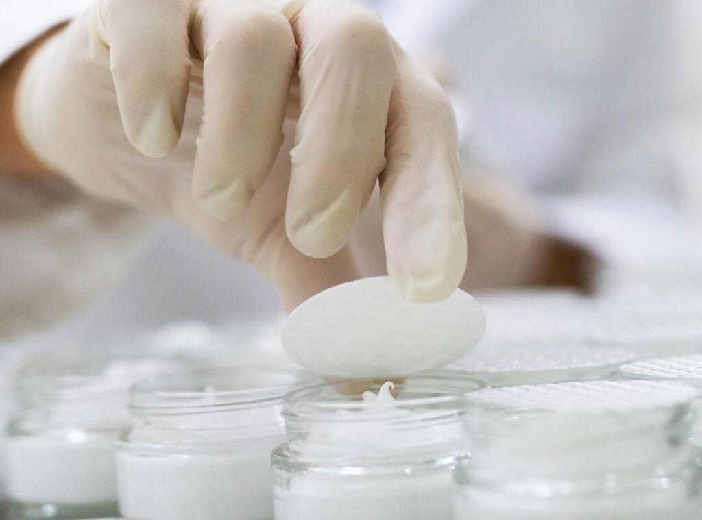 Gloved hand handling a white sample above laboratory containers, illustrating quality assessment of cosmetic raw materials.